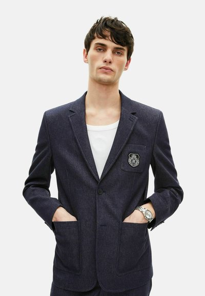 Navyblaues Blazer aus strukturiertem Stoff, mit einem einzelnen Knopfverschluss, zwei Vordertaschen und einem dekorativen Aufnäher auf der Brust.