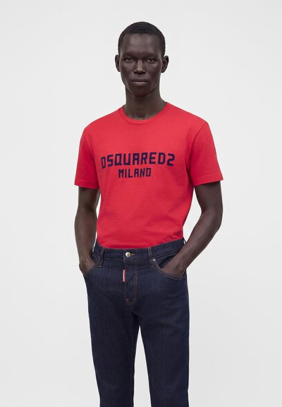 Άνδρας που φοράει κόκκινη μπλούζα με "DSQUARED2 MILANO" σε ναυτικό μπλε κείμενο και σκούρο μπλε τζιν, στέκεται με τα χέρια στις τσέπες μπροστά σε απλό φόντο.