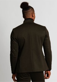 Blazer su misura verde scuro con una superficie liscia, dotato di colletto alto e spalle strutturate, visto da dietro.