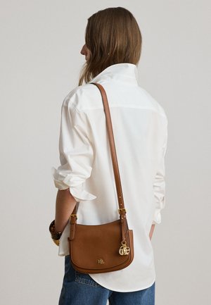 Woman wearing white shirt and blue jeans carrying a brown leather shoulder bag with gold logo charms on a long strap.