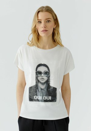 Blond vrouw die een wit t-shirt draagt met een zwart-wit paillettenportret van een persoon met zonnebril en de tekst "OUI OUI."