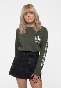 Olive green long-sleeve shirt with white AC/DC logo on the chest and sleeve, paired with a black denim skirt featuring front pockets.