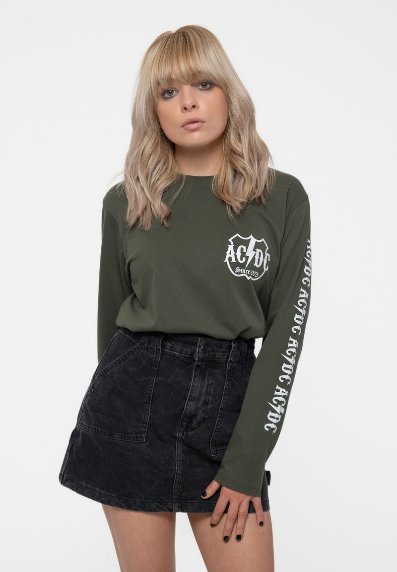 Olive green long-sleeve shirt with white AC/DC logo on the chest and sleeve, paired with a black denim skirt featuring front pockets.