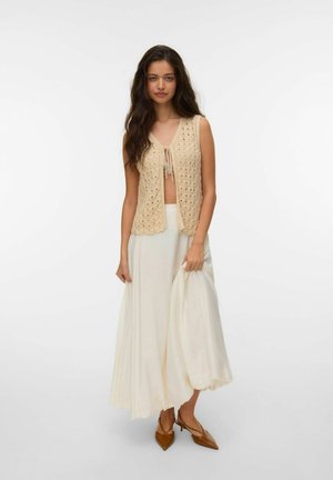 Vrouw met lang donker haar, gekleed in een beige gebreid mouwloos vest, crème vloeiende rok en bruine puntige hakken, staand voor een effen achtergrond.