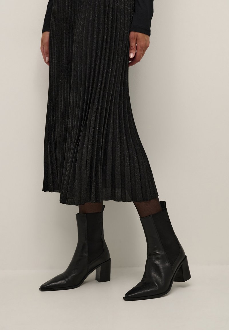 Femme portant une jupe plissée noire mi-longue avec un effet scintillant et des bottines noires à bout pointu avec talons carrés.