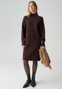 Bruine gebreide jurk met een hoge hals, lange mouwen en een recht model; gecombineerd met een lichtbruine trui. Zwarte enkellaarsjes.