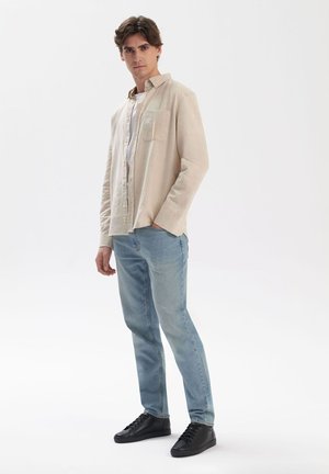 Jasno beżowa koszula z guzikiem i kieszenią na piersi, zestawiona z jasnoniebieskimi dżinsami i czarnymi trampkami, na jednolitym tle.