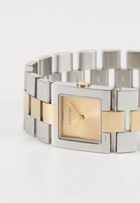 Kandiline kell kuldse näo ja hõbe-kuldse metallkettaga. Omab sileda viimistluse ja minimalistlikku disaini, brändiga "Calvin Klein."