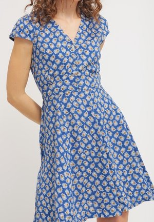 Mujer vestida con un vestido azul con estampado de margaritas blancas y botones en la parte delantera, de pie con un brazo doblado detrás de su espalda.