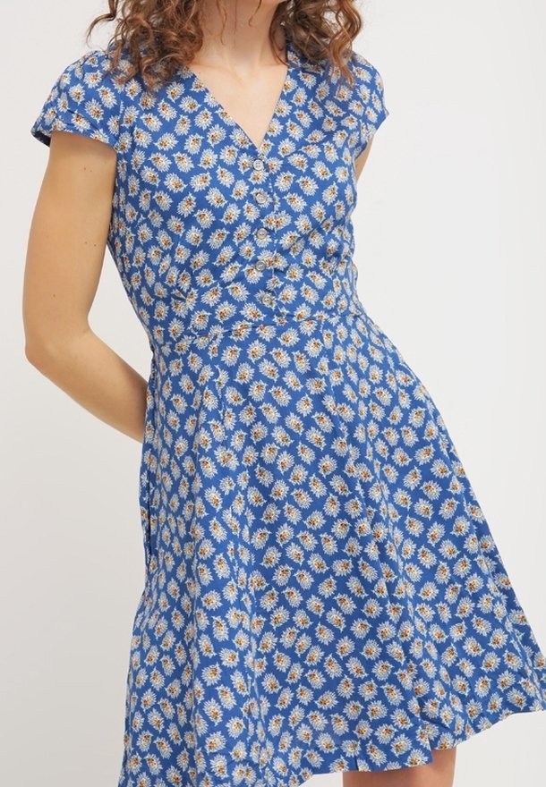 Femme portant une robe bleue avec un imprimé floral de marguerites blanches et des boutons sur le devant, debout avec un bras plié derrière le dos.