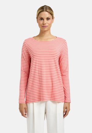 Frau mit blondem Haar, die ein langärmeliges Shirt mit horizontalen Streifen in Rosa und Korallenrot sowie weiße Hosen trägt und vor einem einfarbigen Hintergrund steht.