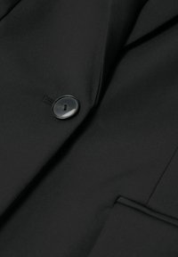 Tissu de blazer noir avec un seul bouton noir fixé au-dessus d'une poche passepoilée sur le côté droit.