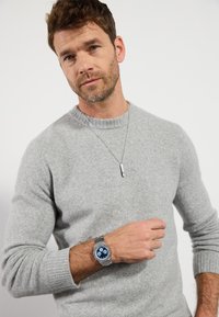 Reloj de pulsera plateado con esfera azul y correa texturizada, llevado en una muñeca. Suéter de lana gris con puños acanalados y cuello redondo.