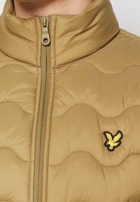 Chaqueta acolchada beige con cuello alto, cremallera central y un logo de águila amarillo y negro en el lado izquierdo del pecho.