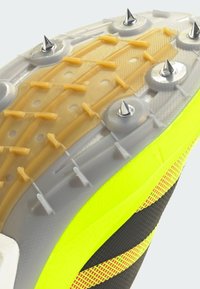 Gros plan sur la semelle d'une chaussure de sport jaune et grise, avec des pointes métalliques et des crampons en caoutchouc pour l'adhérence sur les surfaces de piste.