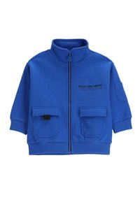 Niebieska kurtka zapinana na zamek z wysokim kołnierzem, dwoma przednimi kieszeniami, ściągaczami przy mankietach oraz nadrukowanym tekstem po lewej stronie. Wykonana z miękkiego, elastycznego materiału.