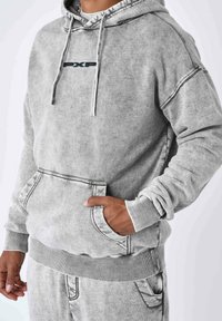 Grijze hoodie van zacht, textuurstof met een kangoeroezak, ribgebreide manchetten en een logopatch in het midden. Contrasterende stikdetails.
