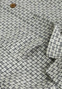 Gewebtes Hemd mit cremefarbenem Hintergrund, das ein geometrisches dunkelblaues Muster, eine Brusttasche und Details mit Holzknöpfen aufweist.