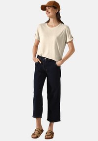Ljusbeige kortärmad skjorta, mörkblå vida jeans och brun keps. Skorna är platta, remmade sandaler med en strukturerad yta.