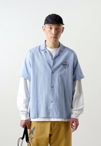 Camicia a maniche corte azzurra con bottoni e logo sul petto, sovrapposta a una maglietta bianca a maniche lunghe, abbinata a pantaloni gialli con ricami.
