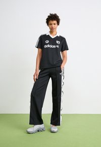 Melnā Adidas sporta krekls ar baltu apkakli un logo, apvienots ar melniem biksēm ar baltām sānu līnijām un plīvojošiem akcentiem. Pelēki sporta apavi.