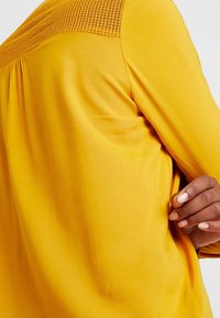 Blusa gialla con yoke testurizzato, tessuto morbido, vestibilità rilassata e dettagli delle cuciture su schiena e lati. Maniche lunghe che si estendono in modo elegante.