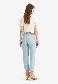 Vaqueros de mezclilla azul claro con un corte relajado, que presentan un parche de cuero marrón con el logo, combinados con una blusa de manga corta color crema y tacones de bloque beige.