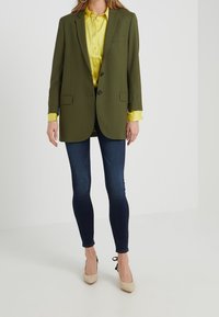 Olivově zelený blazer se dvěma knoflíky, kapsami a přiléhavým střihem, zkombinovaný se světlou žlutou košilí a tmavě modrými úzkými džíny. Béžové vysoké podpatky.