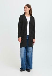 Un long cardigan noir avec une ouverture à l'avant, deux poches avant et une coupe décontractée, porté sur un haut blanc et un pantalon en jean large bleu clair.