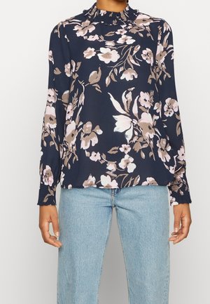 Vrouw draagt een marineblouse met beige en roze bloemenpatronen, een geruite hoge kraag en geplooide manchetten, gecombineerd met lichtblauwe spijkerbroek.