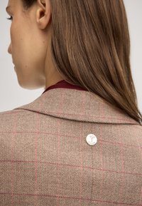 Blazer marrone a motivo houndstooth con check rossi, dotato di colletto a scudetto e di un badge metallico sul retro. Tessuto a texture liscia.