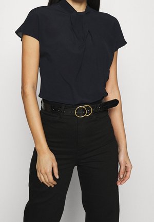 Kobieta w czarnym topie z krótkim rękawem, włożonym w czarne, wysokowalne spodnie z czarnym paskiem ozdobionym dwoma złotymi, okrągłymi klamrami.