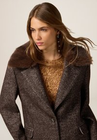 Brauner Fischgrätenmantel mit breitem Kunstfellkragen, Einknopfverschluss und seitlichen Taschen. Darunter ist ein goldfarbener Spitzenoberteil sichtbar.