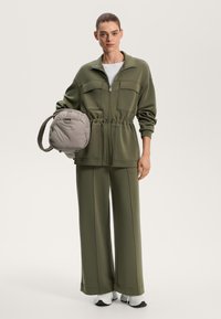 Olive zelena zadrgo jakna s dvema žepoma na prsih, zategnjenim pasom in ujemajočimi širokimi hlačami. Model drži mehak, siv usnjen nahrbtnik.