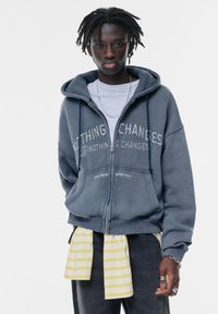 Graue Zip-Hoodie mit dem Text "Nichts ändert sich", lässiger Schnitt, mit vorderen Taschen, über einem weißen Shirt getragen, und einem gelb gestreiften Objekt um die Taille gebunden.
