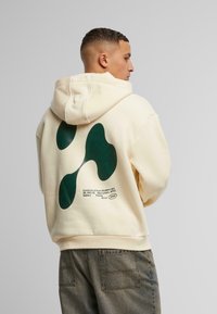 Sweatshirt à capuche crème avec un grand design abstrait vert au dos et du texte imprimé. Style pull surdimensionné avec une coupe décontractée et des poignets côtelés.