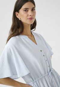 Rochie bleu deschis, cu mânecă scurtă, de tip V, cu închidere cu nasturi și talie ajustabilă cu șnur. Prezintă un material texturat și o croială lejeră.