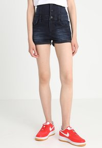 Tmavě modré džínsové kraťasy s vysokým pasem, s předním zapínáním na pět knoflíků, dvěma bočními kapsami a hladkou texturou. V kombinaci s červenými teniskami.