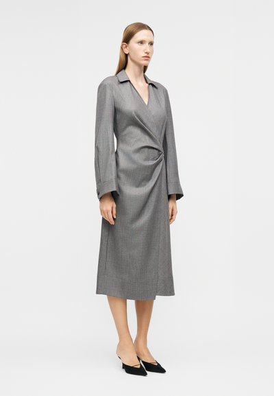 Γυναίκα που φοράει μακρυμάνικο γκρι φόρεμα τύπου wrap με γιακά και μήκος έως τη μέση της γάμπας, συνδυασμένο με μαύρες γόβες με μυτερή μύτη, στέκεται μπροστά σε λευκό φόντο.