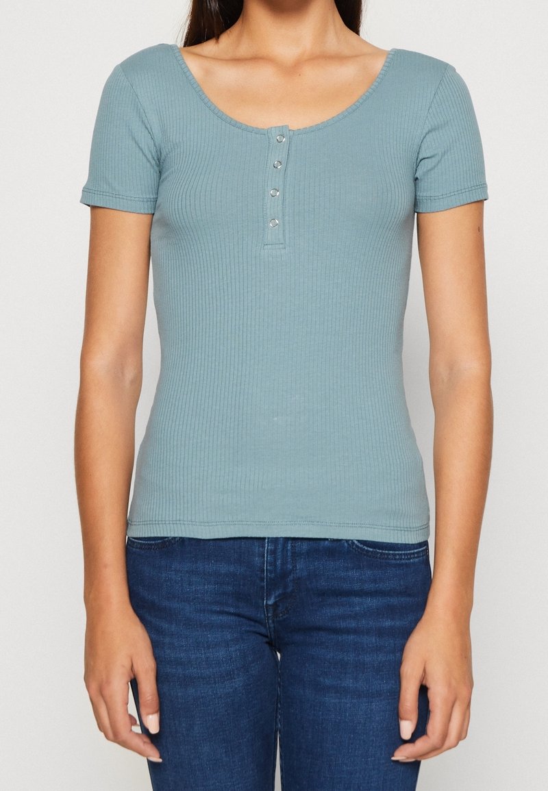 Femme portant un t-shirt bleu côtelé ajusté à manches courtes avec quatre boutons sur le devant, associé à un jean bleu foncé.
