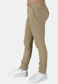 Pantaloni slim-fit beige con una texture liscia, tasche anteriori e un dettaglio con bottone sul retro, abbinati a sneakers bianche.
