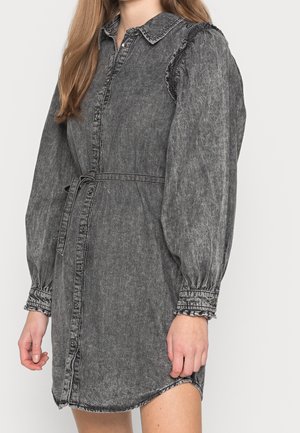 Kvinde iført en falmet sort skjortekjole i denim med lange puffede ærmer, knappet foran, bæltet talje og frynsede detaljer på skuldre og manchetter.