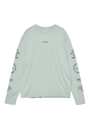 Mężczyzna stoi przodem, ubrany w jasnooliwkową koszulę z długim rękawem z napisem, szare szorty, białe skarpetki i szare trampki na białym tle.