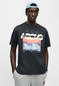 Μαύρο βαμβακερό μπλουζάκι με έγχρωμο γραφικό που απεικονίζει σύννεφα, μεγάλο κείμενο "FLY ABOVE" και λογότυπο της Nike στο κάτω μέρος σε λευκό, φορεμένο με γκρι φόρμες.