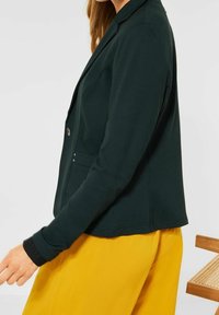 Mörkgrön kavaj med en figursydd design, som har en enkelt knappstängning och sidofickor. Tyget är slätt och kombinerat med gula byxor.