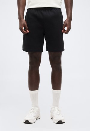 Mann trägt schwarze Shorts, weiße Sportsocken und weiße Turnschuhe, steht vor einem schlichten hellen Hintergrund.