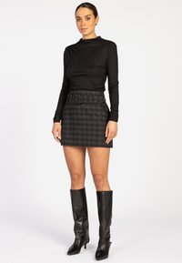 Vrouw in een zwarte langemouwen top, een grijs geruit mini rok met riem en zwarte knie hoge haklaarzen, staande tegen een witte achtergrond.