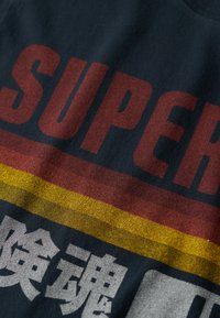 Mørkeblå T-shirt med fed rød "SUPER" tekst og gule og røde striber. Stoffet har en blød tekstur med en let glans.