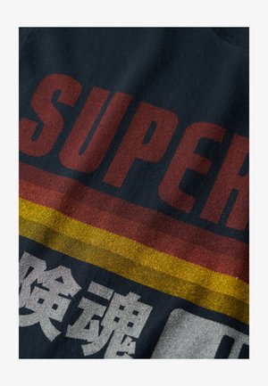 Maglietta blu scuro con il testo rosso in bold "SUPER" e strisce gialle e rosse. Il tessuto ha una texture morbida con una leggera lucentezza.