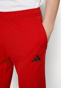 Červené sportovní kalhoty vyrobené z hladkého, elastického materiálu, s černým logem Adidas se třemi pruhy na dolní levé straně a bočními kapsami.